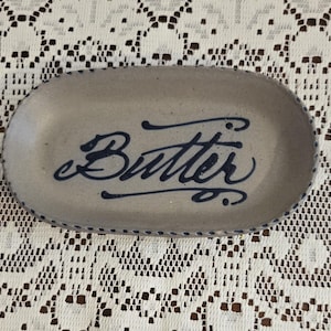 May include: A ceramic butter dish with a light gray base and a navy blue cursive "Butter" inscription. The oval dish has a navy blue dotted border. The dish is on a white lace tablecloth.