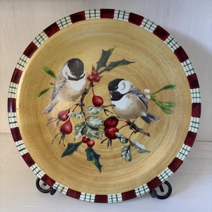 May include: A decorative plate featuring two birds perched on a branch with red berries and green leaves. The plate has a gold-colored center and a red and green checkered border. The plate is displayed on a black metal stand.