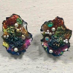 May include: A pair of artisan earrings with a unique, textured design. The earrings feature a mix of colors, including red, green, blue, and pink, with pearl and crystal accents. The irregular shape and intricate details create a statement piece.