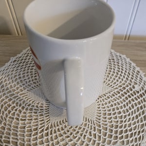 K&ouml;nnte beinhalten: Wei&szlig;er Keramikbecher mit gebogenem Henkel. Der Becher hat ein rot-wei&szlig;es Design an der Seite. Der Becher steht auf einer wei&szlig;en, geh&auml;kelten Untersetzer.