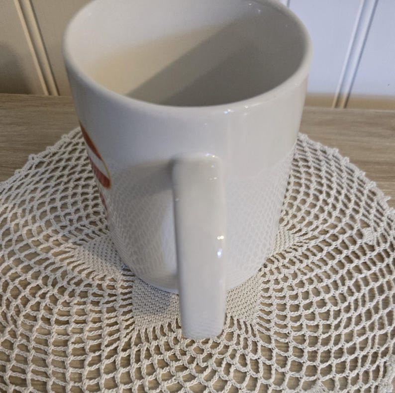 K&ouml;nnte beinhalten: Wei&szlig;er Keramikbecher mit gebogenem Henkel. Der Becher hat ein rot-wei&szlig;es Design an der Seite. Der Becher steht auf einer wei&szlig;en, geh&auml;kelten Untersetzer.