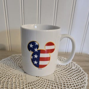 K&ouml;nnte beinhalten: Wei&szlig;e Keramiktasse mit einem Mickey-Mouse-Design in den Farben der amerikanischen Flagge. Die Tasse hat einen gebogenen Henkel und steht auf einer wei&szlig;en Spitzendecke.