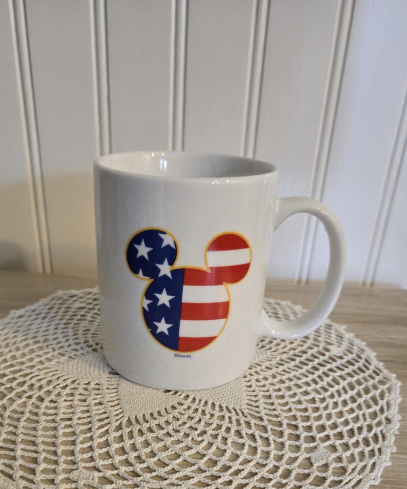 K&ouml;nnte beinhalten: Wei&szlig;e Keramiktasse mit einem Mickey-Mouse-Design in den Farben der amerikanischen Flagge. Die Tasse hat einen gebogenen Henkel und steht auf einer wei&szlig;en Spitzendecke.