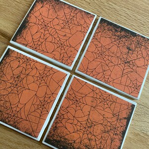 May include: Four square ceramic coasters with a Halloween theme. Each coaster features a black spiderweb design on an orange background with a distressed black border. The coasters have white edges and are arranged on a wooden surface.