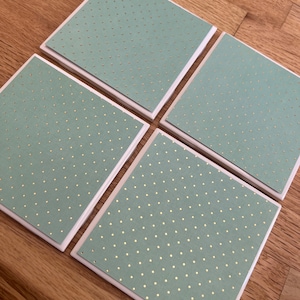 May include: Four square coasters with a light green polka dot design. The coasters have a white border and are arranged on a wooden surface. The polka dots are gold in color.