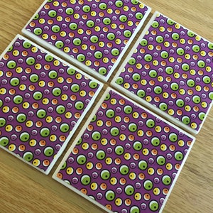 May include: Four square ceramic coasters with a repeating pattern of colorful circles in green, yellow, orange, and purple. The coasters have a white border and are arranged on a wooden surface.
