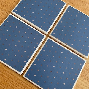 May include: Four square coasters with a navy blue background and a pattern of small, evenly spaced, copper-colored dots. The coasters have a white border and are arranged on a wooden surface.