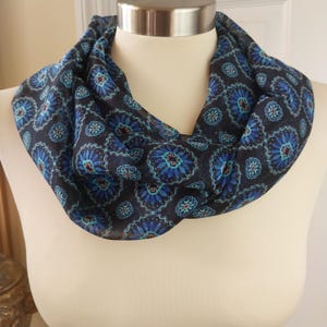 Puede incluir: Una bufanda infinita azul oscuro con un patrón floral repetido en tonos azules y turquesas. La bufanda está drapeada alrededor del cuello de un maniquí, mostrando su diseño y textura. La tela parece ligera y semitransparente.