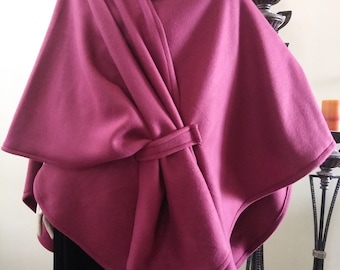 May include: A burgundy cape with a draped design and a knotted closure. The cape is made of a soft material and is worn over a black garment. A beaded necklace is visible at the neckline.