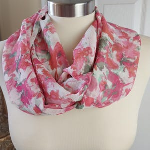 May include: A pink and white floral infinity scarf. The scarf features a repeating pattern of pink, red, and green floral designs on a white background. The scarf is draped around a mannequin, showcasing its circular design.