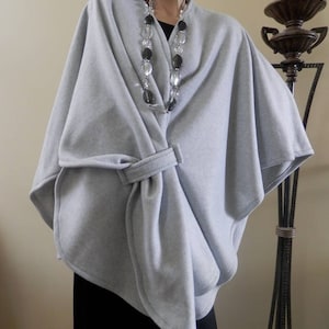 May include: A gray fleece poncho with a belt. The poncho is draped over a mannequin wearing a black dress and a silver and black beaded necklace.
