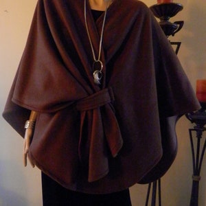 Può includere: Una cappa in pile marrone con chiusura a cravatta. La cappa è indossata sopra una maglia nera e una collana a catena d'argento con un ciondolo decorativo.