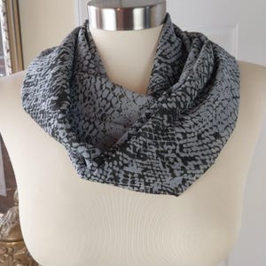 May include: A gray and black infinity scarf with a snake skin pattern. The scarf is draped around a mannequin's neck, showcasing its design and texture. The fabric appears soft and lightweight, suitable for various seasons.