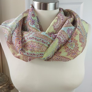 May include: A pastel-coloured infinity scarf with a paisley pattern. The scarf features a repeating design in shades of light green, purple, orange, and blue. The scarf is draped around a mannequin's neck, showcasing its flowing fabric and design.