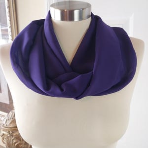 May include: A deep purple infinity scarf displayed on a mannequin. The scarf is made of a soft, flowing fabric and is draped in a figure-eight style. The color is a rich, saturated shade of purple.