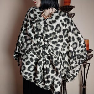 Pode incluir: Um poncho com estampa de leopardo em preto e branco com um design drapeado. O poncho apresenta um decote em V e um detalhe de laço na cintura. O tecido tem uma aparência macia e texturizada. Um colar dourado é visível.