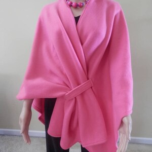 May include: A pink fleece wrap with a tie at the waist. The wrap is worn over a black top and the model is wearing a pink and purple beaded necklace.