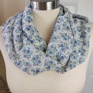May include: An infinity scarf featuring a white base fabric with a repeating floral pattern in shades of dark blue, light blue, and grey. The lightweight material is draped in a circular loop, showcasing the small, dense botanical print throughout.