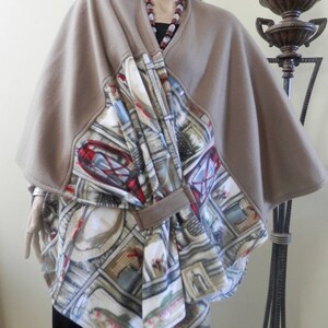 May include: A tan fleece cape with a patterned front panel featuring fishing-themed imagery. The panel includes images of hats, fish, and fishing gear. The cape is styled on a mannequin, showcasing its draped design and overall aesthetic.