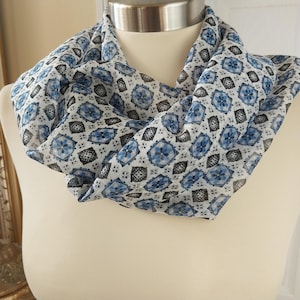 May include: A lightweight, patterned scarf in shades of white, blue, and black. The scarf features a repeating geometric design of blue floral shapes and black diamond-like patterns. The scarf is draped around a mannequin, showcasing its flowy texture.