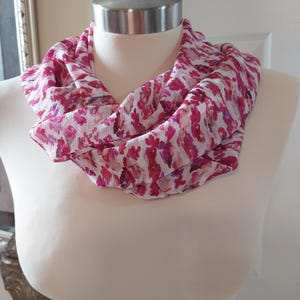 May include: A pink and white infinity scarf with a floral pattern. The scarf is draped around a mannequin's neck, showcasing its design. The fabric appears lightweight and flowing, with a mix of pink and white hues.
