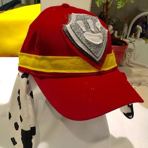 Op de afbeelding: Rode baseballpet met een gele band en een badge met een pootafdruk. De pet heeft wit en zwart gevlekte stof die aan de zijkanten hangt. De badge heeft zilveren glitteraccenten.