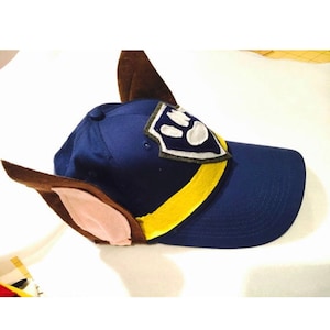 Op de afbeelding: Een marineblauwe baseballcap met bevestigde bruine en beige oren en een gele streep. De cap heeft een badge met een wit pootafdrukontwerp. Een leuk accessoire voor thema-evenementen of dagelijks gebruik.