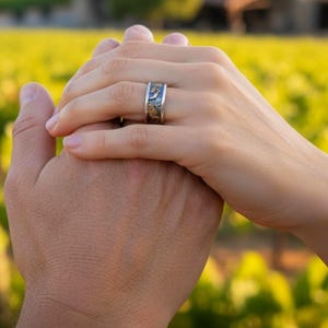 May include: A wide silver band ring featuring a textured, dark oxidized finish with inlaid gold-toned metallic accents. The ring has a flat profile and a polished silver edge, worn on a finger with a vineyard landscape visible in the background.