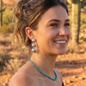 May include: A pair of silver drop earrings featuring three stacked circular discs with engraved spiral patterns, paired with a single-strand turquoise beaded necklace worn with a black off-the-shoulder top.