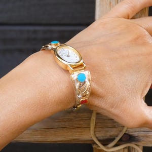 May include: Gold-toned watch with a white face and turquoise accents. The watch band features intricate silver-colored filigree and red details. A classic timepiece with a vintage aesthetic.