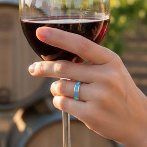 May include: A glass of red wine is held up, with a blue and silver ring on the finger. The background features wine barrels and a vineyard, suggesting a wine-tasting setting. The wine is a deep red color.