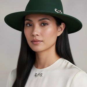May include: A woman wearing a dark green fedora hat with a silver decorative accent. The hat has a wide brim and a matching silver brooch on the white blouse. The woman has long black hair.