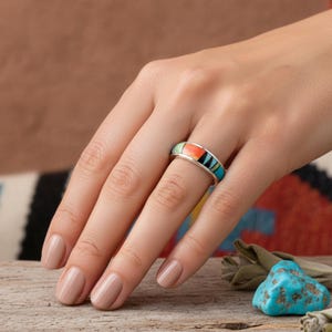 May include: A silver ring with a colorful inlay design featuring turquoise, orange, black, and white stones. The ring is shown on a hand with neutral-toned nail polish. The ring's design is a band style, with a flat top.