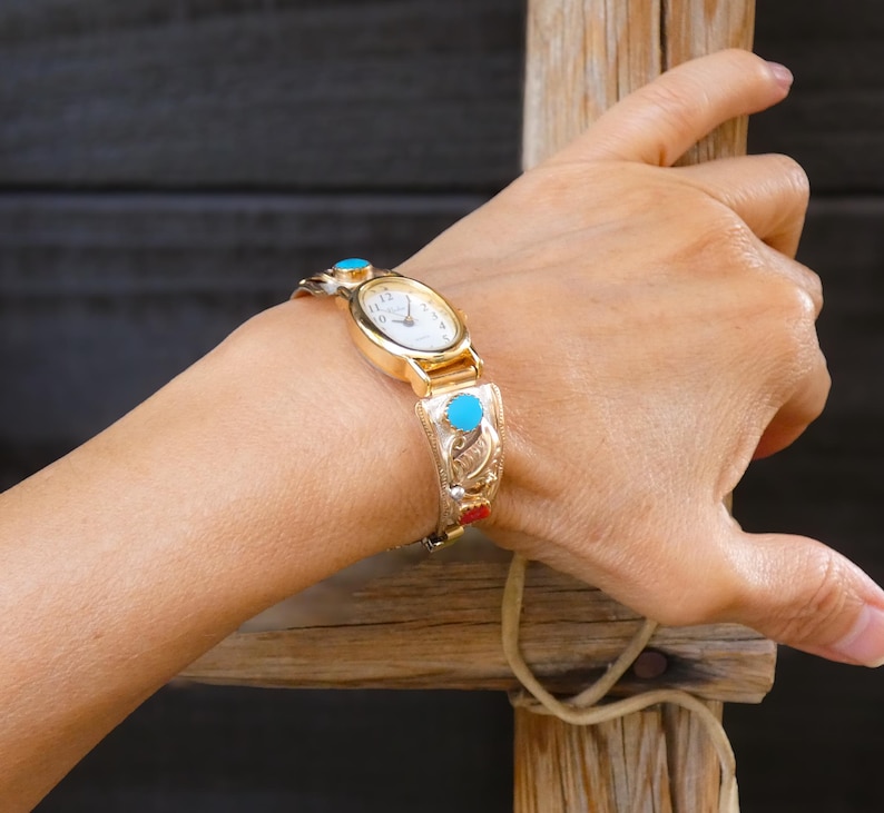 May include: Gold-toned watch with a white face and turquoise accents. The watch band features intricate silver-colored filigree and red details. A classic timepiece with a vintage aesthetic.