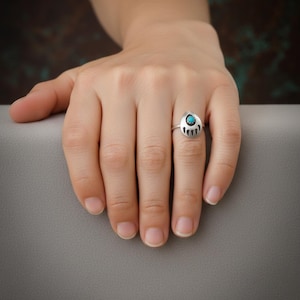 May include: A silver ring with a teardrop-shaped face. The ring features a small, round, turquoise stone at the top and a stylized design at the bottom. The ring is on a finger.
