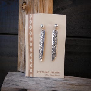 May include: A pair of sterling silver earrings with a geometric design. The earrings are long and rectangular, with a small ball at the top. The card behind the earrings reads "STERLING SILVER" and "The Silver Mesa". The background is a wooden surface.