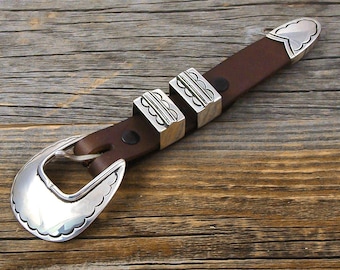 Könnte beinhalten: Ein brauner Ledergürtel mit silberfarbenen Zierakzenten. Der Gürtel hat eine große Schnalle, zwei rechteckige Schlaufen und eine Spitze, alle mit aufwendigen Designs. Der Gürtel liegt auf einem rustikalen Holzgrund.