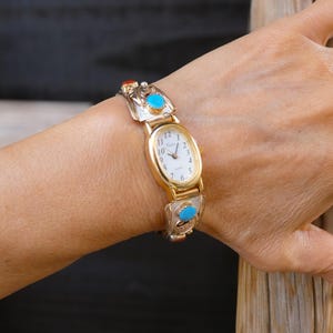May include: Gold-toned wristwatch with an oval face and a white dial. The watch band features silver-toned links with turquoise and coral-colored accents. The watch is worn on a wrist.