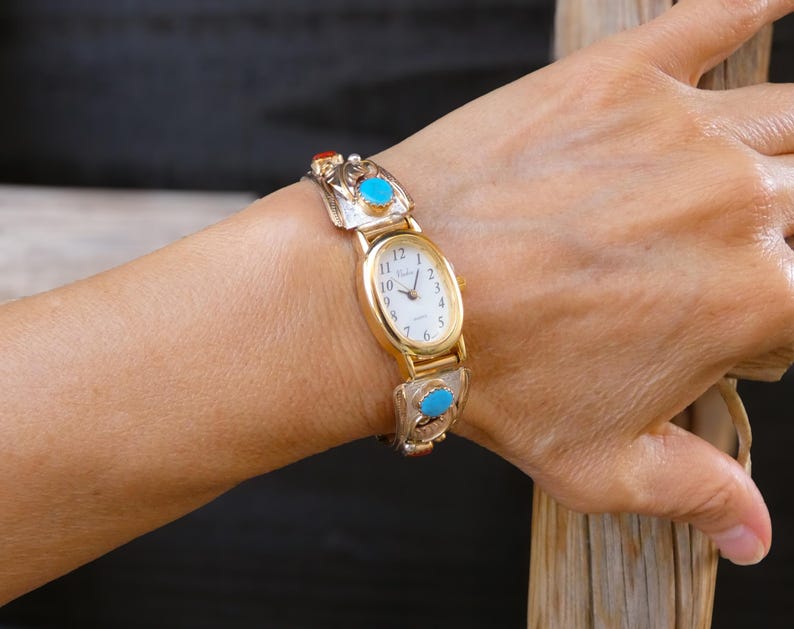 May include: Gold-toned wristwatch with an oval face and a white dial. The watch band features silver-toned links with turquoise and coral-colored accents. The watch is worn on a wrist.