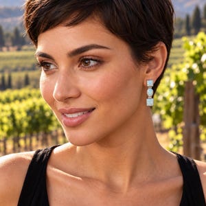 May include: A drop earring featuring a vertical arrangement of three stones set in metal. The design consists of a square stone at the top, an oval stone in the center, and a square stone at the bottom, all displaying an iridescent white finish.
