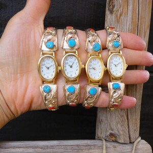 May include: Four gold-toned watches with oval faces and white dials are displayed. Each watch features turquoise and red accents, with ornate, engraved bands. The watches are held up against a dark background and a wooden post.