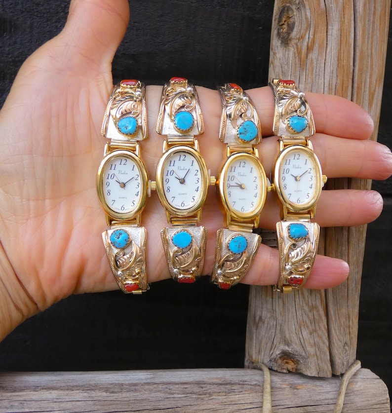 May include: Four gold-toned watches with oval faces and white dials are displayed. Each watch features turquoise and red accents, with ornate, engraved bands. The watches are held up against a dark background and a wooden post.