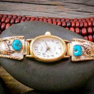 May include: A gold-toned watch with an oval face and a white dial, featuring the brand name "Nadia". The watch band has silver and gold accents with turquoise stones. The watch is displayed on a dark gray stone.