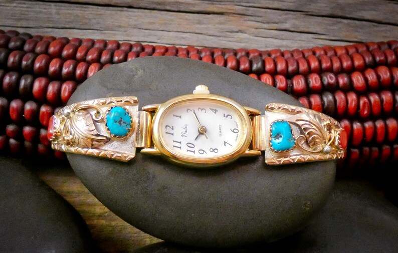 May include: A gold-toned watch with an oval face and a white dial, featuring the brand name "Nadia". The watch band has silver and gold accents with turquoise stones. The watch is displayed on a dark gray stone.