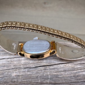 May include: A gold-toned wristwatch with an oval face and a flexible band. The watch is set against a weathered wooden surface, showcasing its metallic sheen and intricate design. The band is made of small, rectangular links.