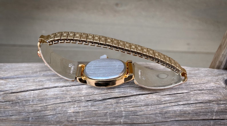 May include: A gold-toned wristwatch with an oval face and a flexible band. The watch is set against a weathered wooden surface, showcasing its metallic sheen and intricate design. The band is made of small, rectangular links.