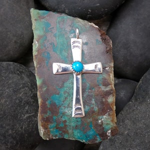 May include: A silver cross pendant with a turquoise stone center. The cross has decorative sunburst patterns on each arm and a loop for a chain. The pendant is displayed on a textured, multi-colored stone.