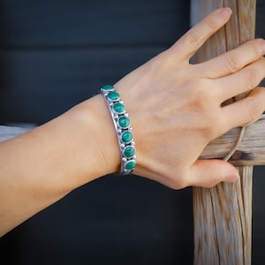 May include: A silver cuff bracelet featuring seven circular, vivid green stones. The bracelet is displayed on a wrist, highlighting the contrast between the polished metal and the stones' colour. The design is simple but elegant.