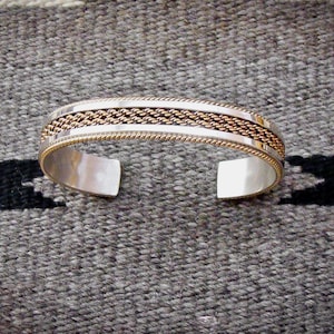 May include: Silver cuff bracelet with a gold-toned woven center and gold-toned trim. The open-ended bracelet is displayed on a gray woven surface. The bracelet's design features a combination of silver and gold tones.