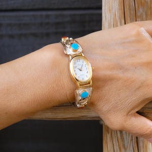 May include: Gold-toned wristwatch with an oval face and a white dial. The watch band features silver and turquoise accents. The watch is worn on a wrist, showcasing the design and craftsmanship of the timepiece.